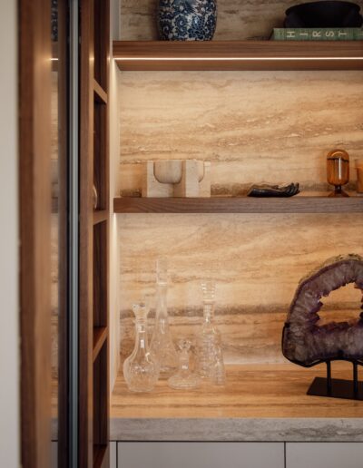 Wooden shelves with crystal decanters, a geode, books, a blue and white vase, and small decorative objects against a beige stone wall.