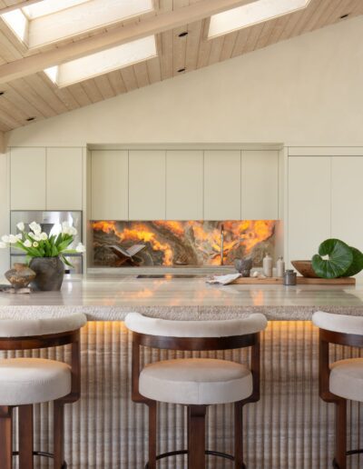 Modern kitchen with a large marble island, five upholstered stools, neutral cabinetry, and a backlit stone backsplash. Natural light enters from skylights in the angled wooden ceiling.