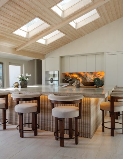 Modern kitchen with a large island, cushioned bar stools, built-in appliances, under-cabinet lighting, stone backsplash, and skylights in a wood-paneled ceiling.