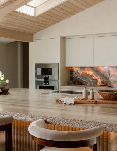 Modern kitchen with a large island, neutral tones, built-in appliances, stone backsplash with dramatic veining, and skylight overhead providing natural light.