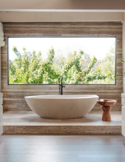 Modern bathroom with a freestanding oval bathtub centered beneath a large window, flanked by double vanities and surrounded by natural wood and stone accents.
