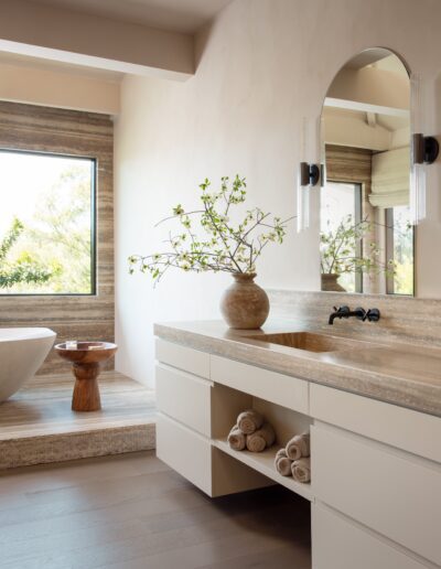 Modern bathroom with a stone countertop, built-in sink, large mirror, freestanding tub, wooden stool, and a vase with green branches on the vanity.