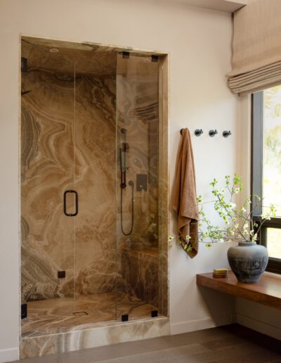 A marble walk-in shower with glass doors, a wall-mounted towel and hooks, a wooden bench, and a ceramic vase with green branches next to a window.