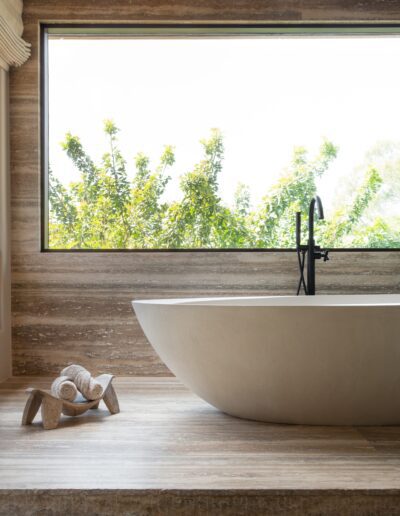 A modern bathroom features a freestanding oval bathtub next to a large window with a tree view, and a small stool holding rolled towels.