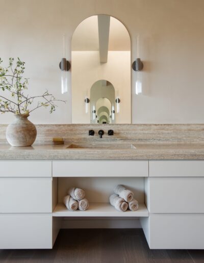 Modern bathroom vanity with a stone countertop, built-in sink, rolled towels on a shelf, a potted plant, and an arched mirror reflecting a series of similar mirrors.