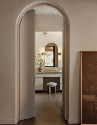 A view through an arched doorway into a minimalist bathroom with a vanity, a round stool, a vase with branches, and a framed artwork leaning against the wall.