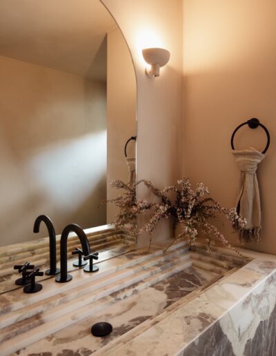 A marble sink with black fixtures, a large mirror, a wall sconce, a towel on a black ring, and a vase with branches in a softly lit bathroom.